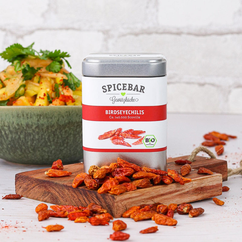 Birdseyechilis-Dose von Spicebar auf Holzbrett, umgeben von getrockneten roten Chilis, mit frischem asiatischem Gericht im Hintergrund.