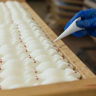 Blick auf eine Hand mit blauen Handschuhen, die rote Zuckeraugen mit Spritztülle auf Schaumzuckermäuse aufträgt.