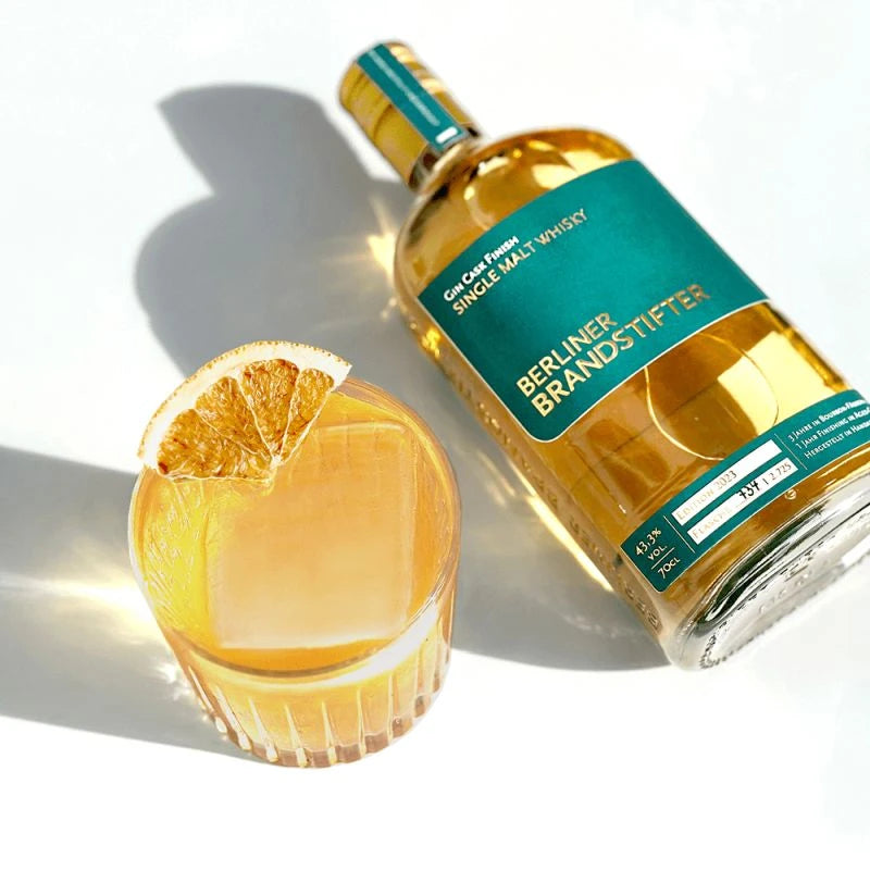 Stilvolle Szene mit einer Flasche Berliner Brandstifter Single Malt Whisky neben einem befĂŒllten Tumblerglas mit groĂem EiswĂŒrfel und Orangenzeste.