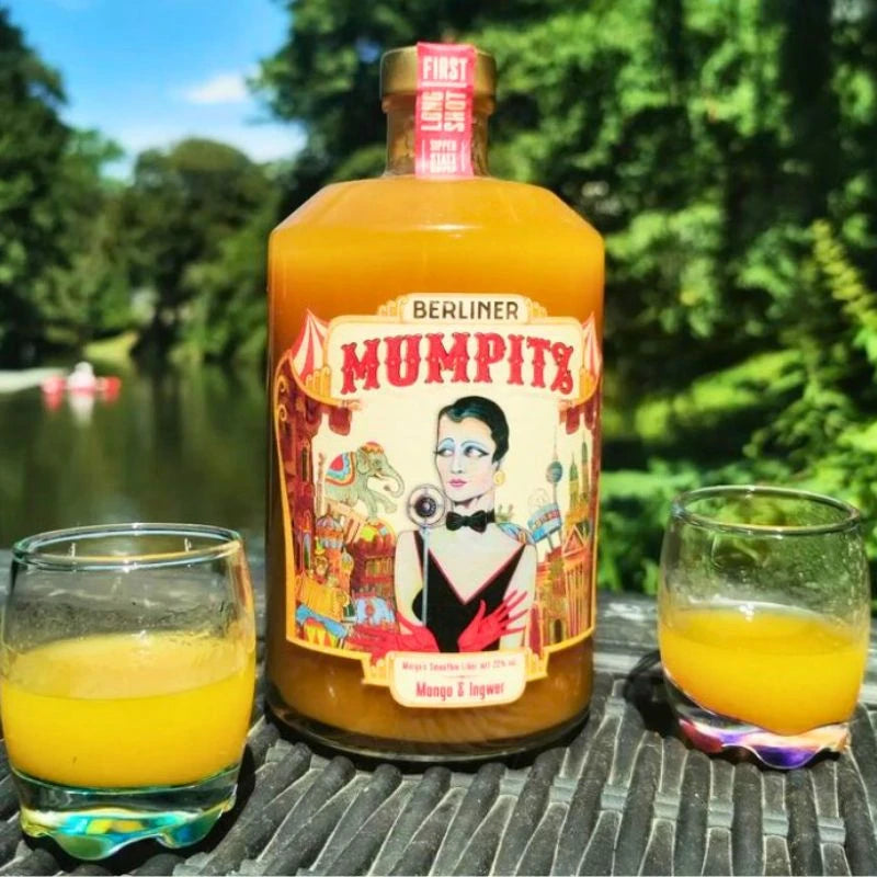 Szene im Freien mit Berliner Mumpitz Mango & Ingwer auf einem Tisch, flankiert von zwei Gläsern mit dem fruchtig-würzigen Likör; sommerliche Stimmung am Wasser.