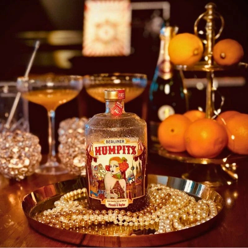 Berliner Mumpitz Pfirsich & Hopfen Flasche auf Tablett mit Perlenkette, umgeben von Orangen, Cocktailgläsern und Champagnerflasche in stilvoller Bar-Atmosphäre.