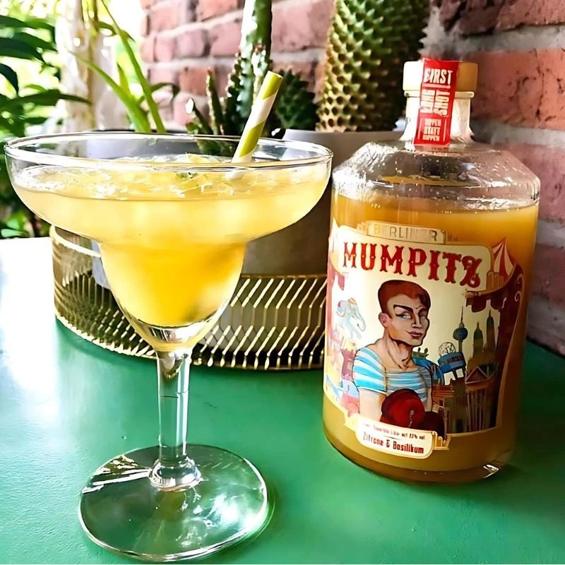 Berliner Mumpitz Zitrone & Basilikum Likörflasche auf grünem Tisch mit gefülltem Cocktailglas – sommerliches Szene mit Pflanzen im Hintergrund.