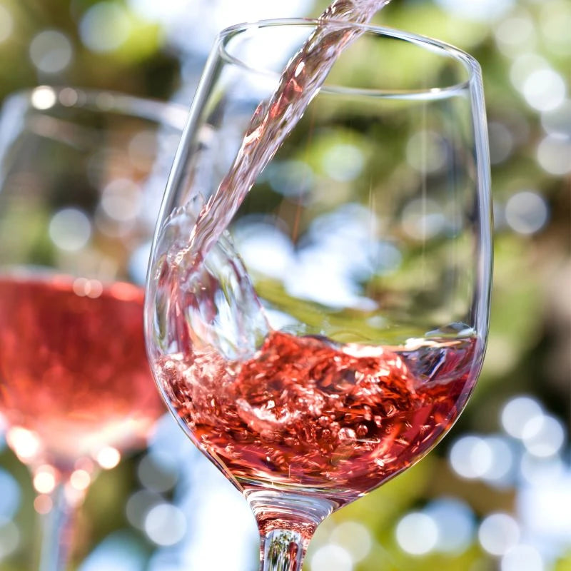 Rosé Secco wird in ein Weinglas gegossen – der Perlwein leuchtet im Sonnenlicht, umgeben von sommerlich-grünem Bokeh. Sinnbild für Berliner Lebensfreude, Leichtigkeit und bewussten Genuss in Bio-Qualität.