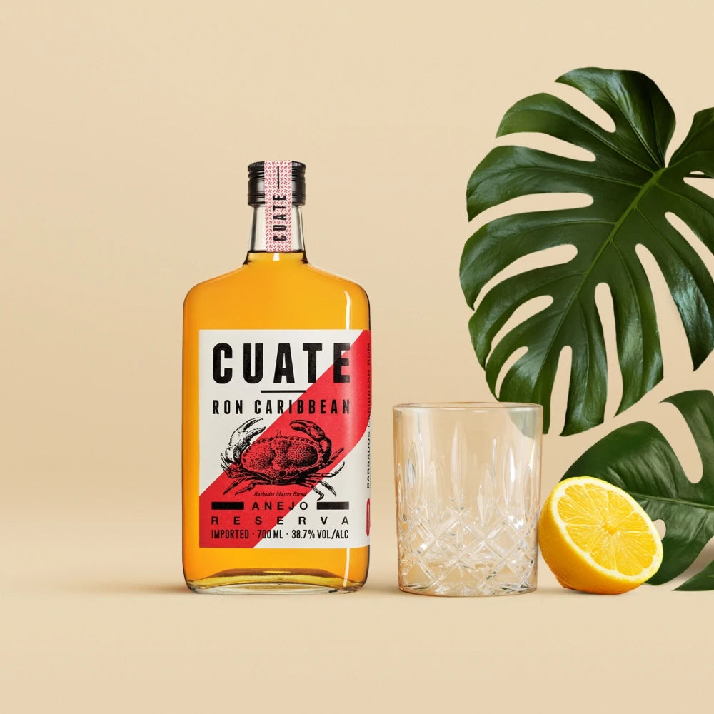 CUATE Añejo Reserva Rum neben Tumbler-Glas, halber Zitrone und Monstera-Blatt auf beigem Hintergrund – karibisches Setting.