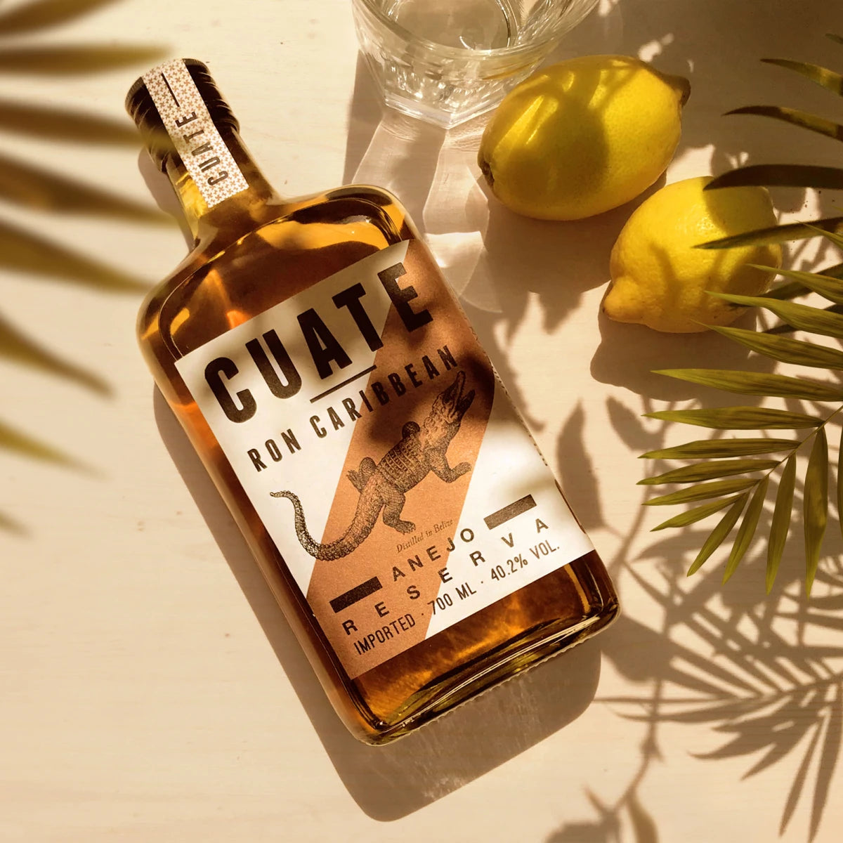 CUATE Rum 06 Añejo Reserva – Rumflasche mit Alligator-Etikett liegend im Sonnenlicht, dekoriert mit Zitronen und Palmenblättern.