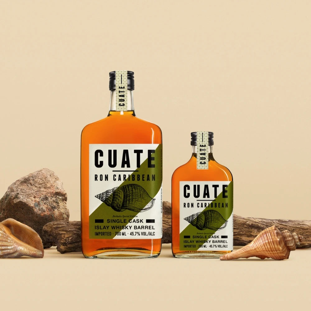 CUATE Ron Caribbean Islay Whisky Barrel in 0,7 l- und 0,2 l-Flaschen nebeneinander auf Naturkulisse mit Muscheln, Holz und Steinen – karibisches Design trifft Berliner Rum-Ästhetik.