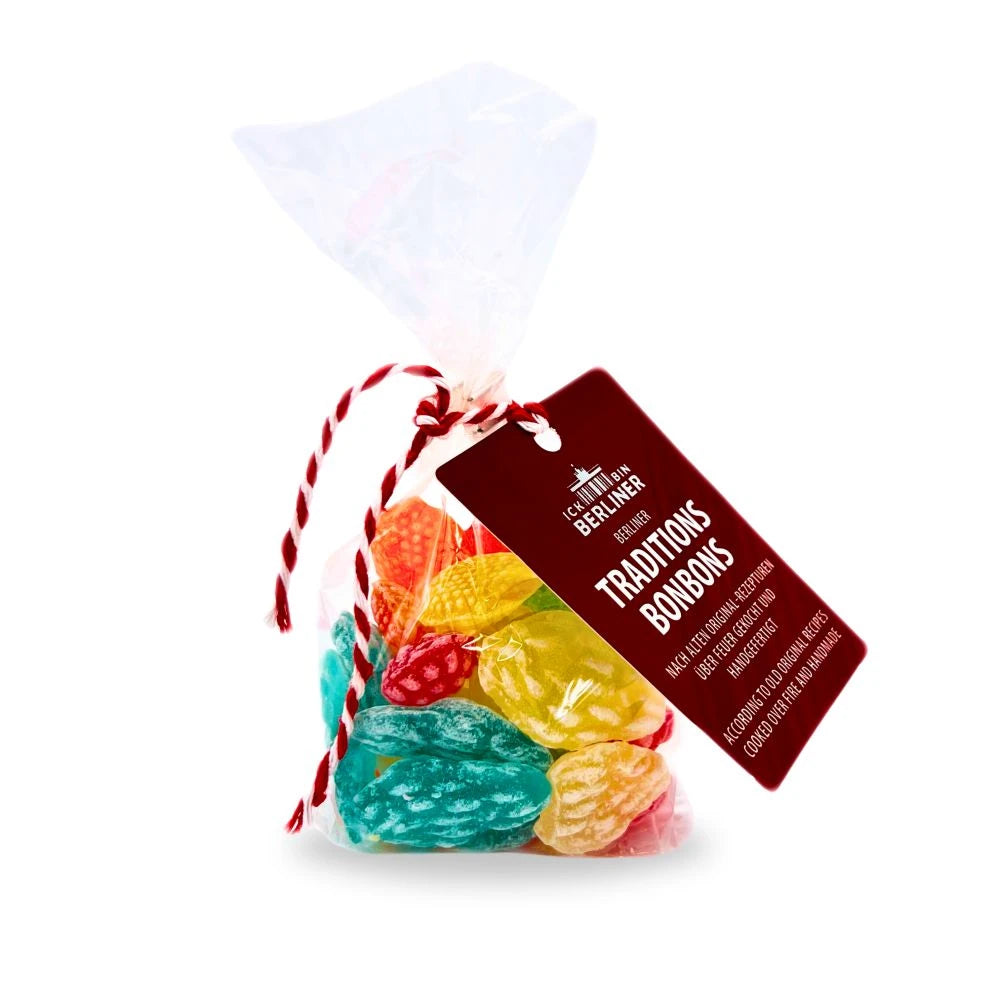 Ein durchsichtiger Cellophanbeutel, gefüllt mit handgemachten Berliner Traditionsbonbons in fruchtigen Farben. Die Bonbons sind in klassischen Formen wie Beeren und Blättern gestaltet. Am Beutel hängt ein rotes Etikett mit der Aufschrift ‚ICK BIN BERLINER – Traditionsbonbons – nach alten Original-Rezepturen über Feuer gekocht und handgefertigt‘.