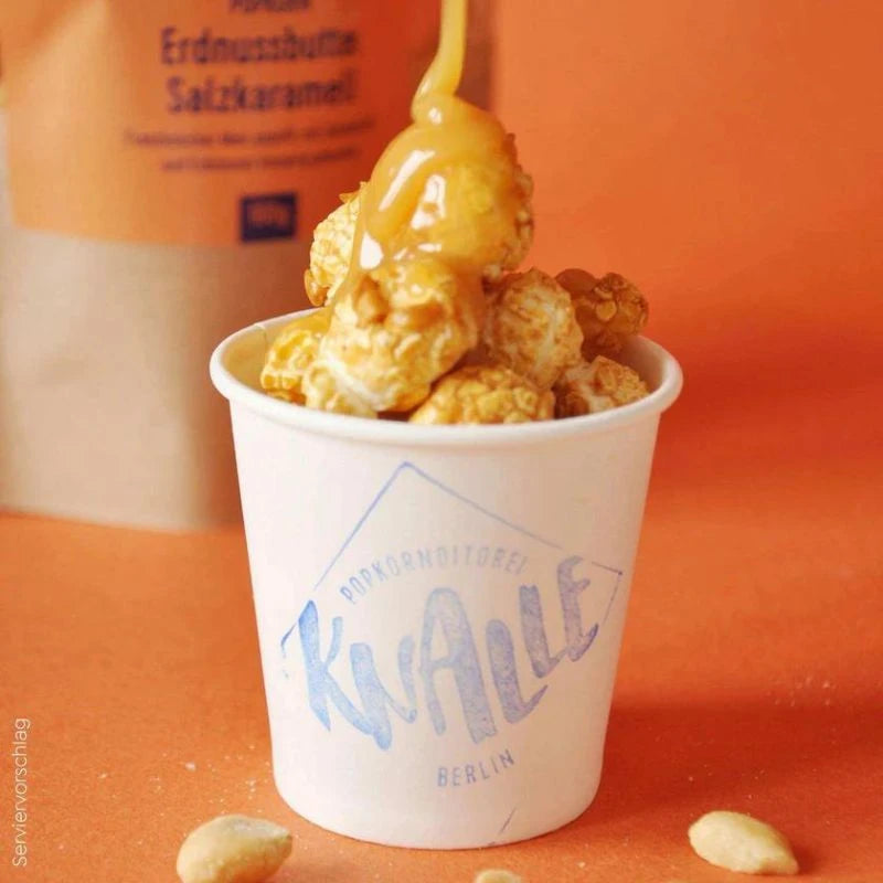 Serviervorschlag: Popcorn Peanut Butter Salted Caramel in weißem Becher, übergossen mit flüssigem Karamell. Präsentiert auf orangem Hintergrund mit Erdnüssen als Deko.