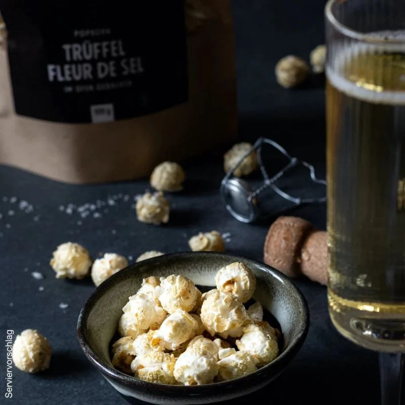 Knalle Popcorn Trüffel Fleur de Sel in Keramikschale, daneben Champagner, Korken und Salzflocken – stilvolle Snack-Inszenierung für besondere Anlässe.