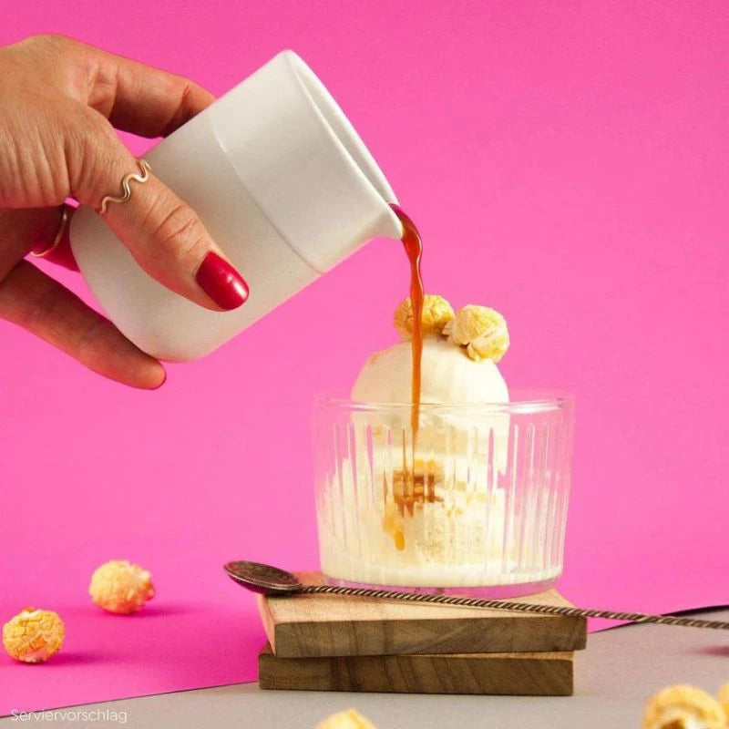 Serviervorschlag: Butterkaramell-Popcorn als Topping auf Vanilleeis, dazu wird Karamellsauce aus einem weißen Becher gegossen – vor pinkem Hintergrund.