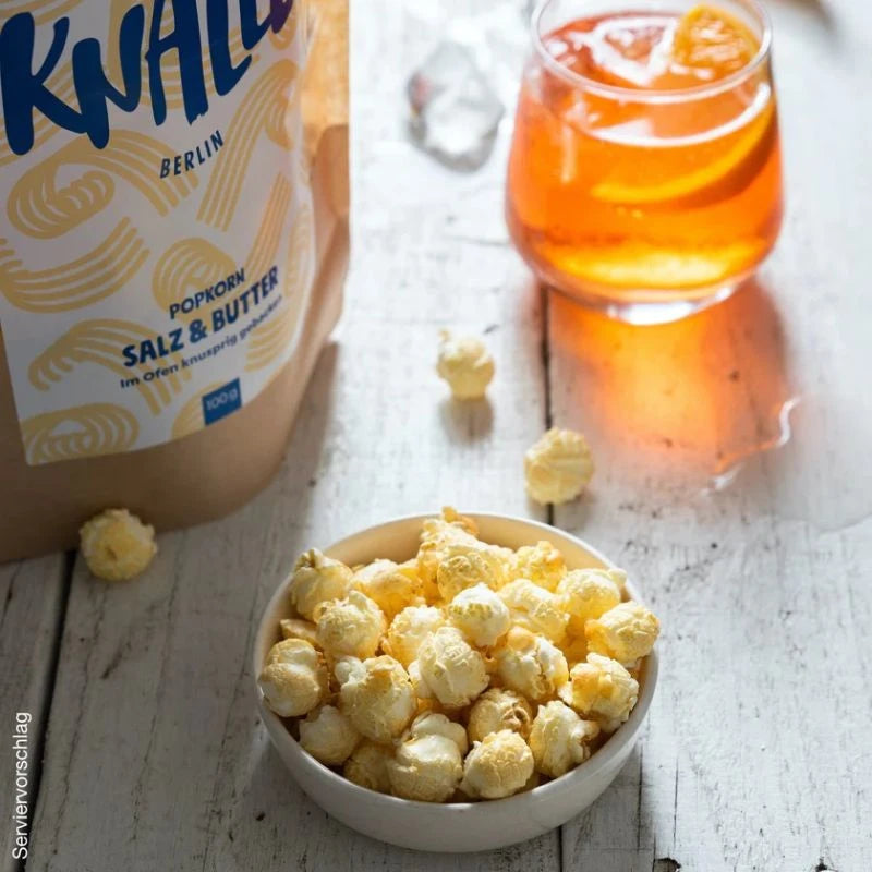 Knalle Popcorn Salz & Butter in weißer Schale auf rustikalem Holztisch, daneben ein Glas mit Orangenlimonade – perfekter Snackmoment mit Berliner Manufakturqualität.