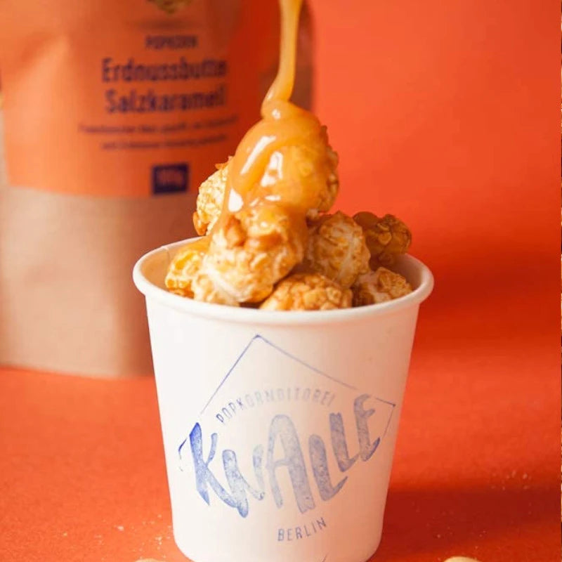 Knalle Popcorn Erdnussbutter Salzkaramell im weißen Becher, übergossen mit flüssigem Karamell – vor orangem Hintergrund. Berliner Popkornditorei zum Naschen und Veredeln.