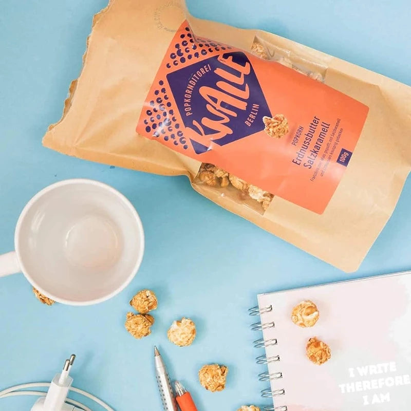 Geöffnete Tüte Knalle Erdnussbutter Salzkaramell Popcorn auf Schreibtisch mit Tasse, Stift und Notizbuch. Perfekter Berliner Snack für kreative Pausen.