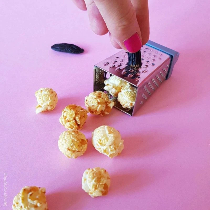 Serviervorschlag: Popcorn mit Kokos und Tonkabohne, daneben eine Mini-Reibe mit geriebener Tonkabohne auf pinkem Hintergrund – kreative Darstellung des Berliner Snacks.