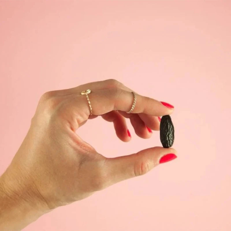 Tonkabohne wird zwischen zwei Fingern gehalten – Nahaufnahme vor rosa Hintergrund. Die aromatische Zutat verleiht dem veganen Knalle Popcorn seinen charakteristischen, vanillig-nussigen Geschmack.