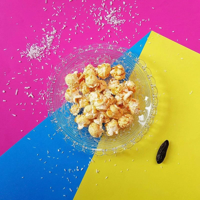 Popcorn mit Kokos in einer gläsernen Schale, präsentiert auf farbenfrohem Untergrund in Pink, Blau und Gelb. Dekoriert mit Kokosraspeln und ganzer Tonkabohne.
