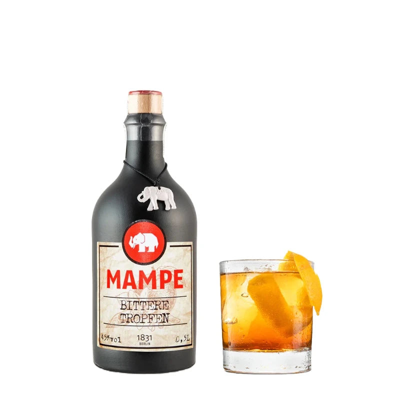 Mampe Bittere Tropfen Flasche neben einem Tumblerglas mit bernsteinfarbenem Drink auf Eis, garniert mit Orangenzeste. Der Drink wirkt aromatisch, herb und charaktervoll.