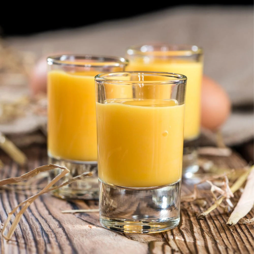 Drei Shotgläser gefüllt mit cremigem, gelbem Eierlikör auf einem rustikalen Holztisch mit Strohdeko. Die Szene wirkt ländlich, natürlich und einladend.