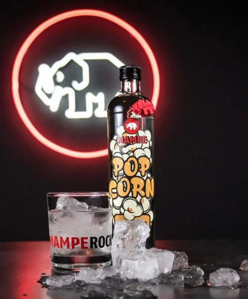 Mampe Popcorn Likör Flasche vor einem Neon-Elefantenlogo, inszeniert mit Tumblerglas, Eiswürfeln und Markenbranding „MAMPE ROCKT“. Die Szene wirkt urban, cool und nightlife-inspiriert.