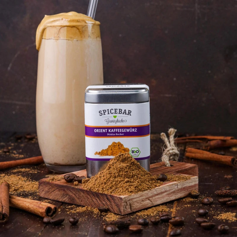 Stimmungsvolles Arrangement mit einer Dose Spicebar Orient Kaffeegewürz, Zimtstangen, Kaffeebohnen und einem Dalgona-Kaffee auf dunklem Holzuntergrund.