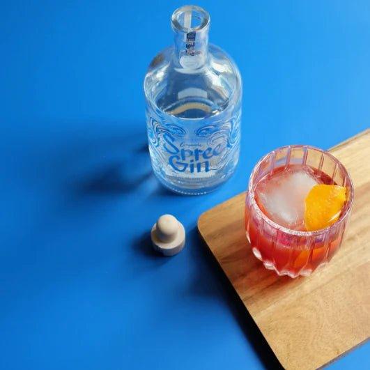 Spree Gin Berlin Dry Gin – leere 0,5 l Flasche mit Holzverschluss neben einem frisch gemixten Cocktail mit Eis und Orangenzeste auf Holzbrett, stilvoll inszeniert auf blauem Hintergrund.
