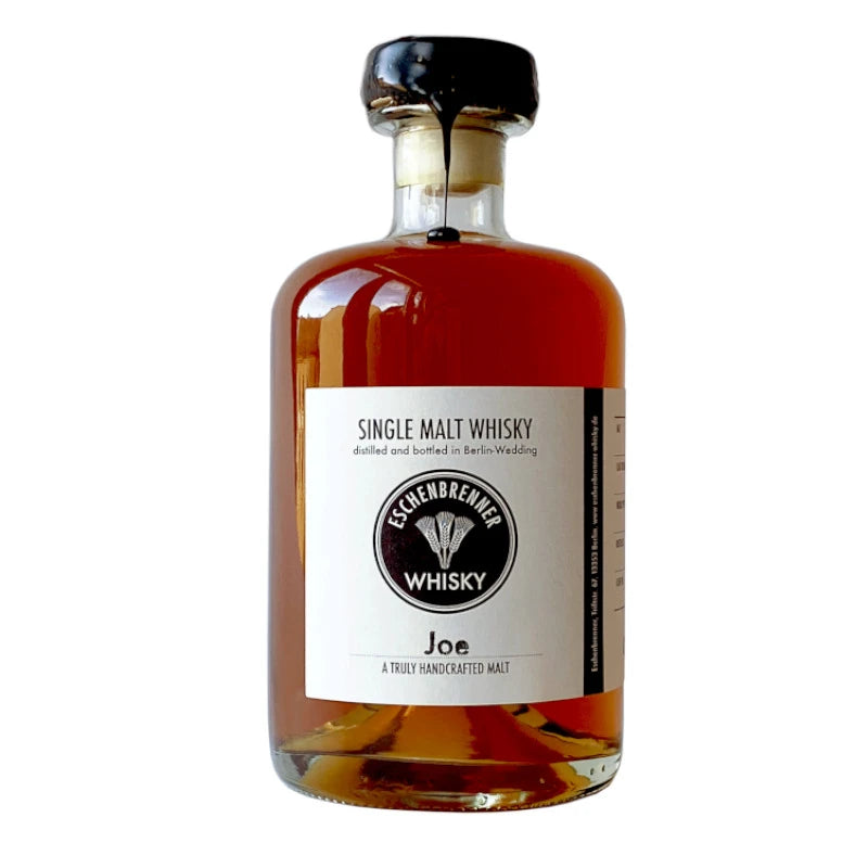 Eschenbrenner Single Malt Whisky „Joe“ in klarer 0,5 l-Flasche mit schwarzem Wachsverschluss. Weißes Etikett mit Logo, Aufschrift „Single Malt Whisky – distilled and bottled in Berlin-Wedding“ und dem Namen „Joe – A truly handcrafted malt“.