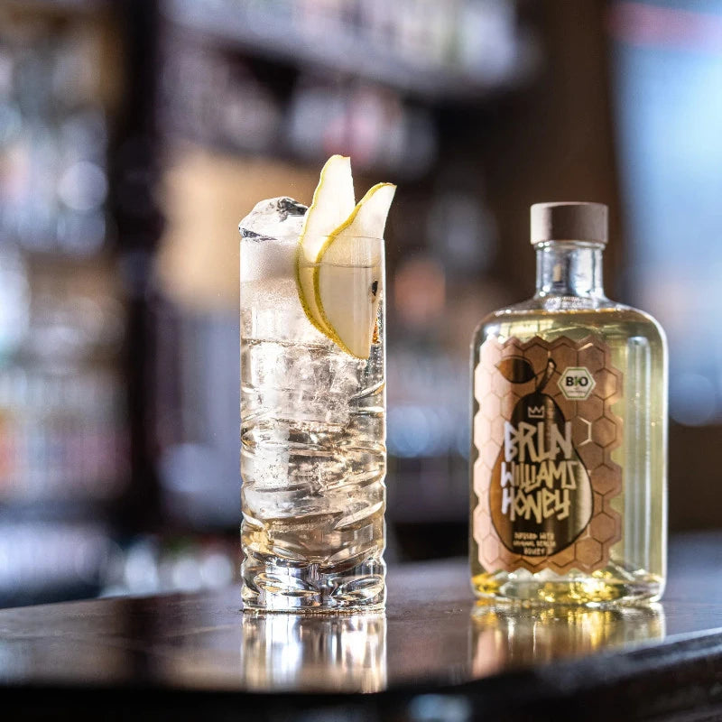 Grote BIO Williams Honey 0,5 l neben einem hohen Glas mit eisgekühltem Williams-Cocktail und Birnenscheiben, goldgelber Bio-Likör aus Berlin in Bar-Atmosphäre