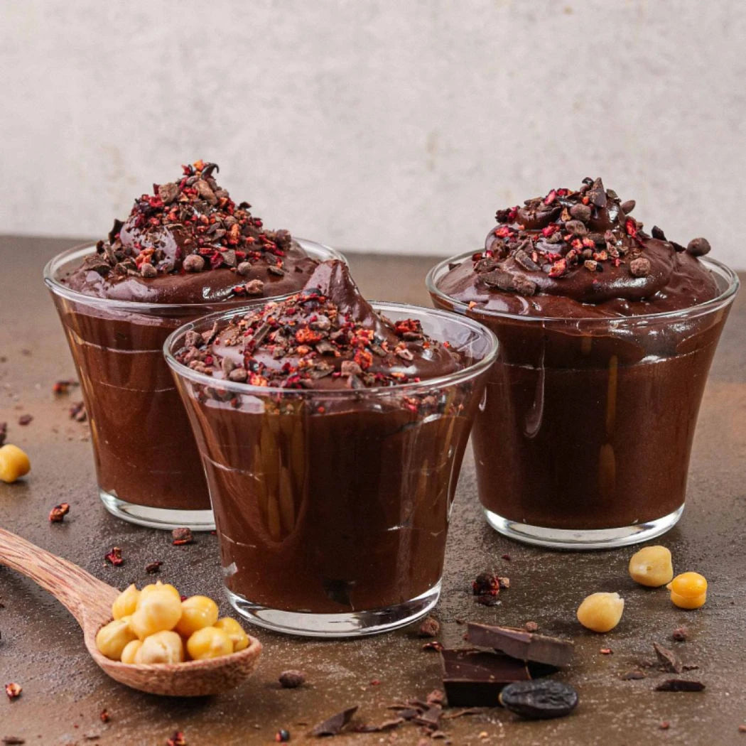 Drei Gläser mit cremigem, dunklem Schokodessert, dekoriert mit Gewürzen und Kakaosplittern, daneben Kichererbsen und Zartbitterschokolade.